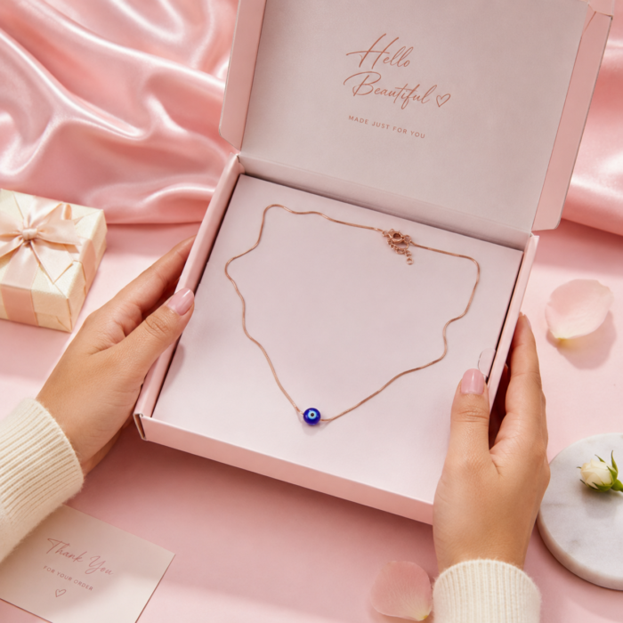 Hands opening a pink gift box revealing a delicate rose gold-toned chain necklace with a blue evil eye pendant.